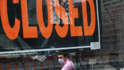 Window into a business with a closed sign and a worker with a mask on