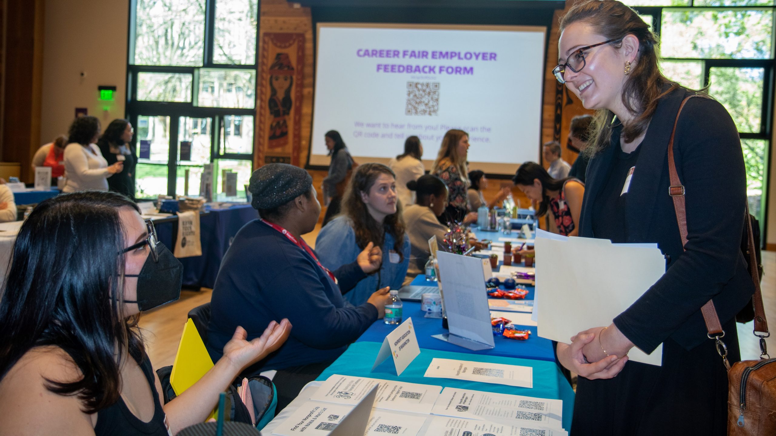 A student holding a folder of papers talks to a potential employer seated at an information table