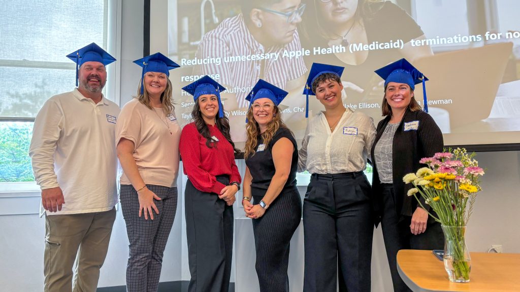 A group of six people wearing blue graduation caps in front of a presentation screen, with a vase of flowers in the foreground.