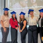 A group of six people wearing blue graduation caps in front of a presentation screen, with a vase of flowers in the foreground.