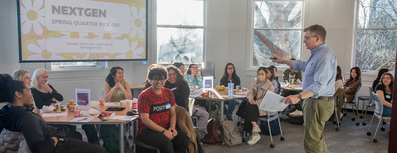 NextGen students sit at tables smiling at each other while listening to an instructor who is speaking to the group. A projected slide on the screen behind everyone reads ``NextGen Spring Quarter Kick Off``