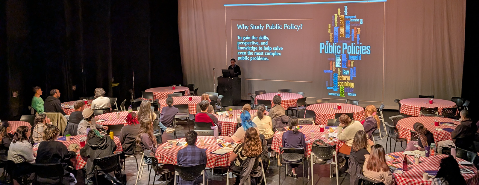 Policy Camp students sit at round tables that are covered with red and white checkered tablecloths. A projected slide is on the screen at the front of the room and reads ``why study public policy?`` and features a word map that includes the words: public policies, law, reform, benefit, government, economic, business, social, solve, and judicial