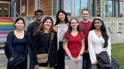 A group of students pose in front of a building with rainbow painted steps and the words "Kenmore City Hall" visible in the background